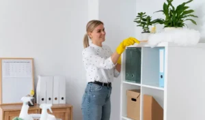 Woman in white top, blue jeans and yellow gloves cleaning cabinets with a white furry duster.