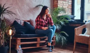 Woman in red check shirt and blue jeans sitting on a wooden couch and looking towards window.