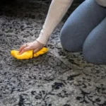 Woman in white t-shirt and jeans holding a spray bottle cleaning carpet with a yellow brush.