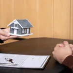 Midsection of person holding small house and contract with keys on table