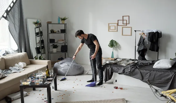 Young man sweeping floor at home after party bottles, crumps and poker chips on floor