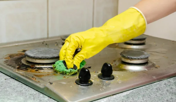 Cleaning dirty gas stove from grease, food leftovers deposits. Woman's hand in protective glove with sponge rag